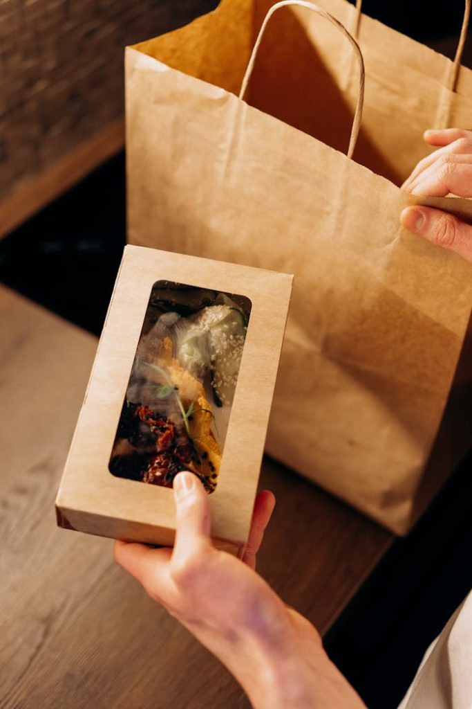 Hands holding a cardboard food container next to a paper bag, suggesting eco-friendly delivery.