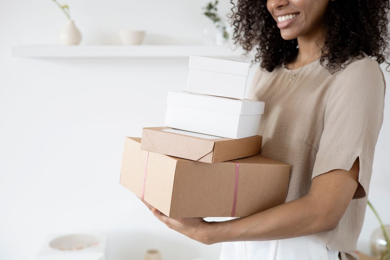 Woman with curly hair carrying stack of packaging boxes in a bright room.